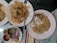 A spread of Indian cuisine with a large serving of biryani garnished with herbs on a white plate, alongside another plate with a partially eaten piece of chicken and rice. There is also a plate containing sliced onions and lemon, with three metal containers holding different sauces or chutneys.