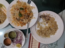 A spread of Indian cuisine with a large serving of biryani garnished with herbs on a white plate, alongside another plate with a partially eaten piece of chicken and rice. There is also a plate containing sliced onions and lemon, with three metal containers holding different sauces or chutneys.
