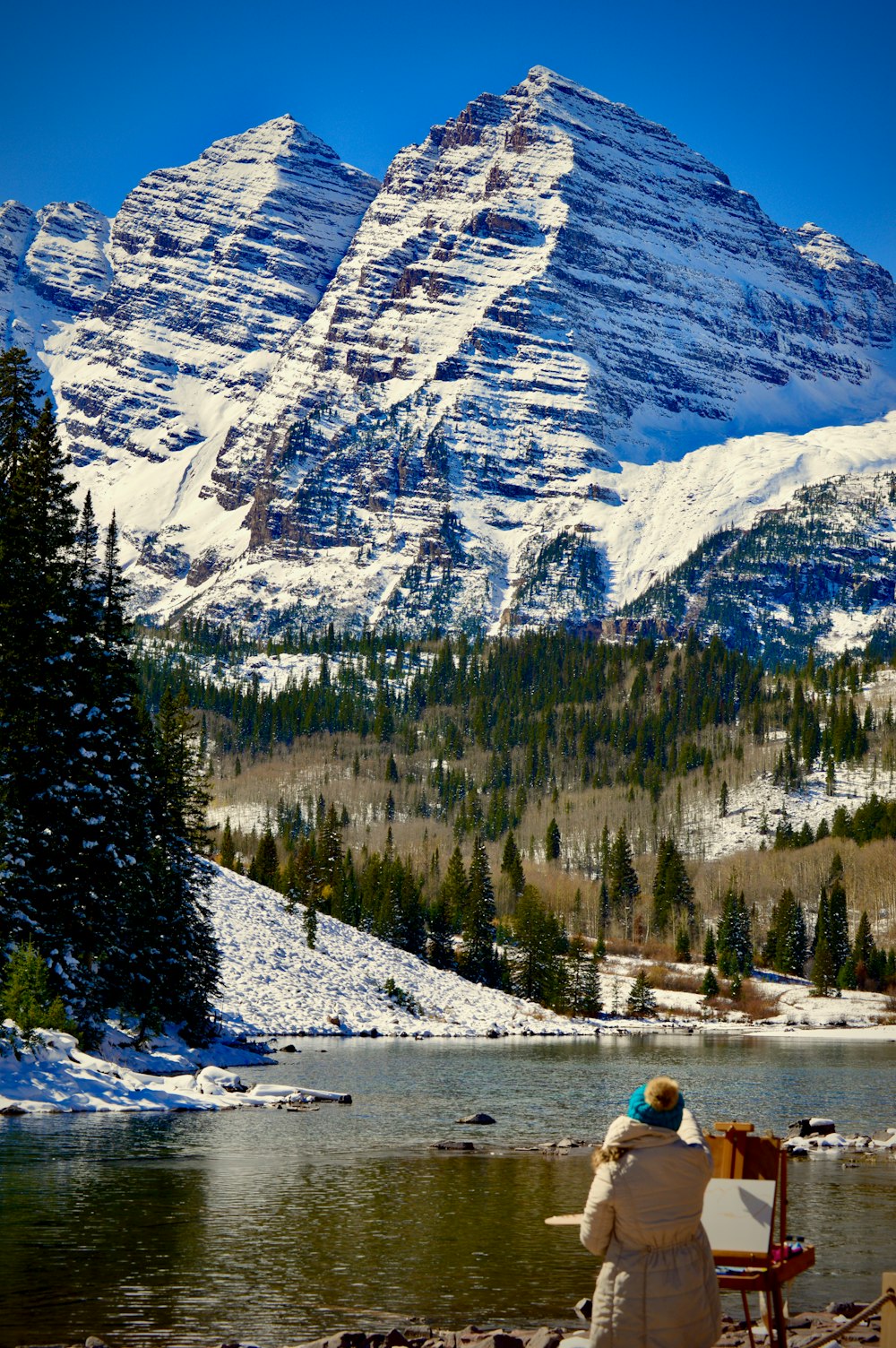 woman wearing brown coat standing near easel board beside lake viewing mountain covered with snow during daytime