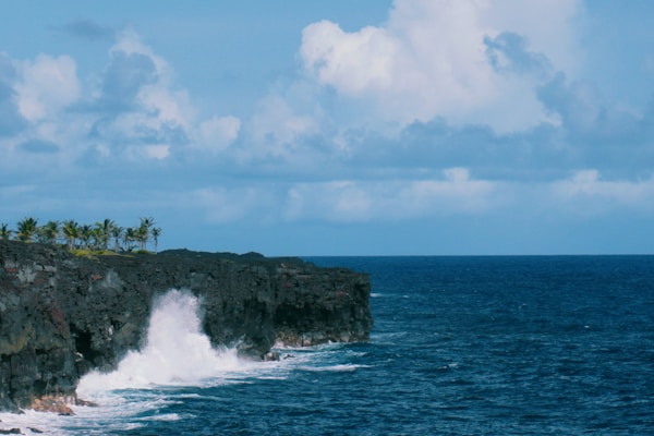 Waves crash against rugged cliffs under a partly cloudy sky, with a few palm trees visible in the distance. The vibrant blue of the sea contrasts with the dark, rocky cliff face.