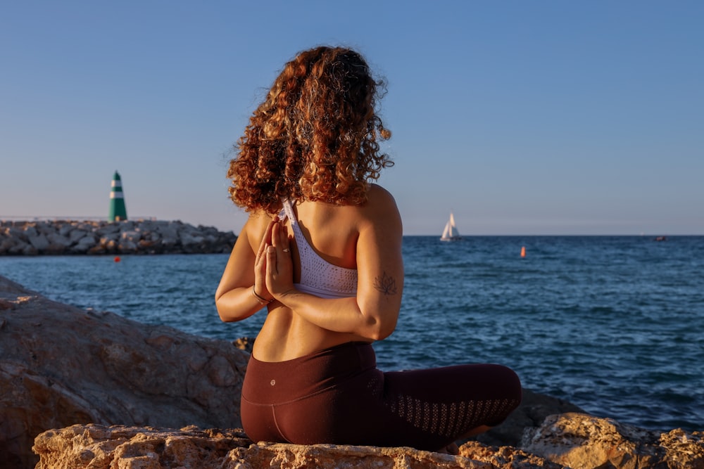 woman sitting while doing yoga on rock viewing body of water during daytime