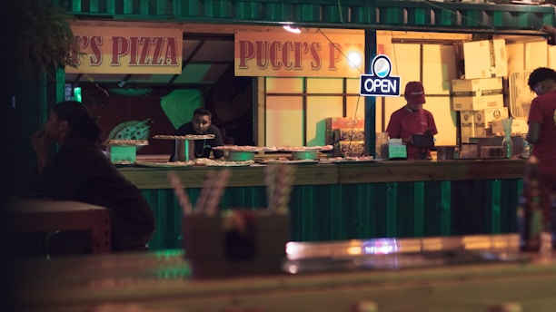 A dimly lit pizza stall with a sign reading 'Pucci's Pizza' and an 'OPEN' neon light. Several pizzas are displayed on the counter. Two employees, one wearing a red hat, are behind the counter. A customer sits at a nearby table, seemingly waiting.