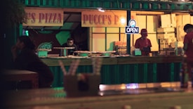 A dimly lit pizza stall with a sign reading 'Pucci's Pizza' and an 'OPEN' neon light. Several pizzas are displayed on the counter. Two employees, one wearing a red hat, are behind the counter. A customer sits at a nearby table, seemingly waiting.