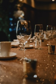 A dimly lit, elegant table setting featuring several wine glasses and white cups with saucers. The table is covered with a shimmering cloth, and gold-colored votive candle holders are placed alongside. The background is blurred, with soft, warm lighting adding to the sophisticated ambiance.