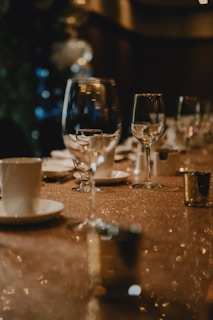 A dimly lit, elegant table setting featuring several wine glasses and white cups with saucers. The table is covered with a shimmering cloth, and gold-colored votive candle holders are placed alongside. The background is blurred, with soft, warm lighting adding to the sophisticated ambiance.
