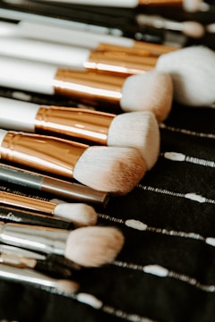 A collection of makeup brushes with soft, fluffy bristles and metallic handles are neatly arranged on a dark fabric surface. The bristles are light-colored and appear to have been crafted for applying cosmetics.