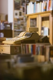 An old-fashioned cash register or adding machine is situated on a counter surrounded by shelves filled with books. The background is slightly blurred, suggesting a cozy, retro environment that might be a bookstore or library.