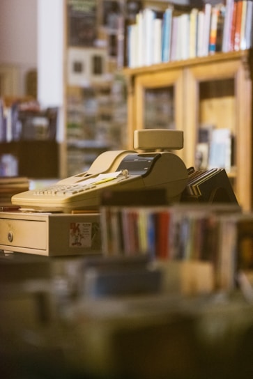 An old-fashioned cash register or adding machine is situated on a counter surrounded by shelves filled with books. The background is slightly blurred, suggesting a cozy, retro environment that might be a bookstore or library.
