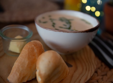 A bowl of creamy soup garnished with herbs sits on a wooden tray. Next to it are two pieces of bread and a small glass dish containing butter. The background features blurred lights and objects, adding a cozy and warm ambiance.