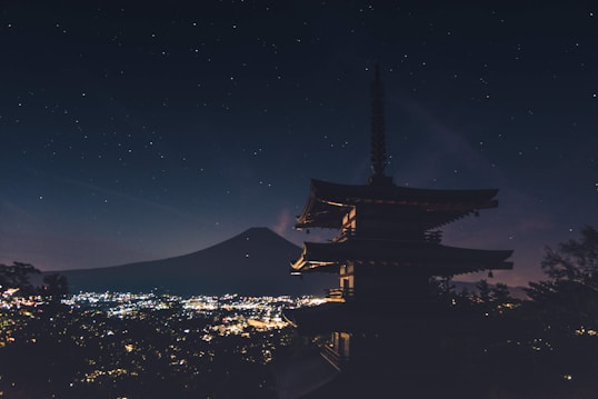 A silhouette of a traditional Japanese pagoda stands prominently against a starry night sky. In the background, the outline of Mount Fuji is visible, with twinkling city lights sprawling below, creating a serene and majestic atmosphere.