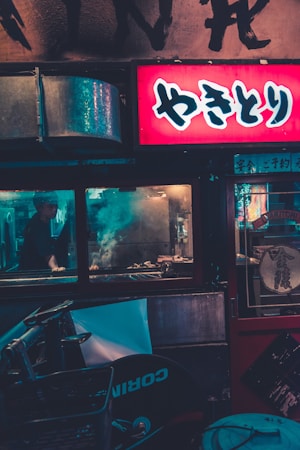 A street food stall with a prominent, illuminated red sign featuring Japanese characters. The interior is dimly lit with smoke rising from a grill where food is being prepared. A person is partially visible behind the window, tending to the grill. The area outside the stall is cluttered with various objects, including parts of a bicycle.