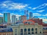 A historic train station with a prominent sign reading 'Union Station Travel by Train' is set against a backdrop of a modern city skyline. The skyline features various high-rise buildings, with a clear blue sky overhead adorned with wispy clouds.