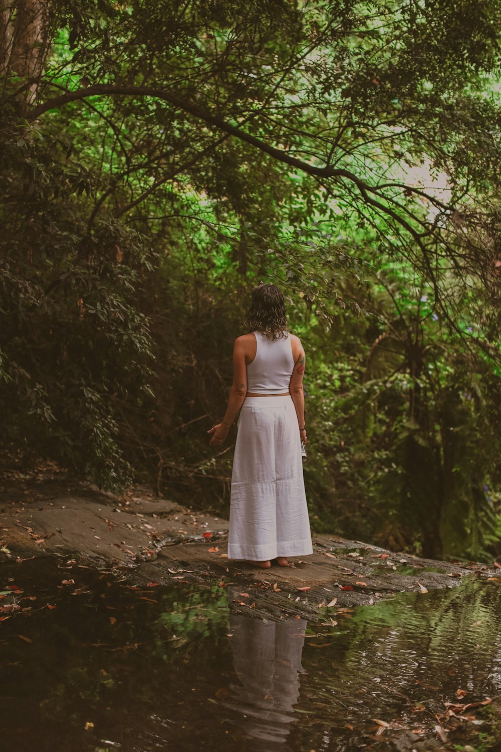 woman in white tank top and white pants standing on boulder near body of water