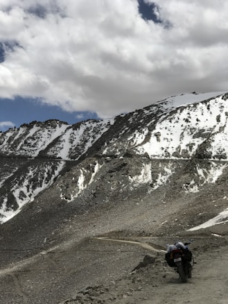 A rugged mountain landscape with patches of snow covering parts of the rocky terrain. A single motorcycle with luggage is parked on a narrow dirt path winding through the mountains. The sky is partly cloudy, casting shadows on the slopes.