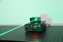 A green laser system placed on a perforated metallic surface. The system consists of a main driver unit labeled 'DPSSL DRIVER FA' and an emitting unit. The front panel of the driver unit has a digital readout displaying the value '1325', buttons, and indicator lights. A visible green laser beam is emitted from the top unit, projecting onto a wall in the background, creating a green spot and a faint shadow of the apparatus.