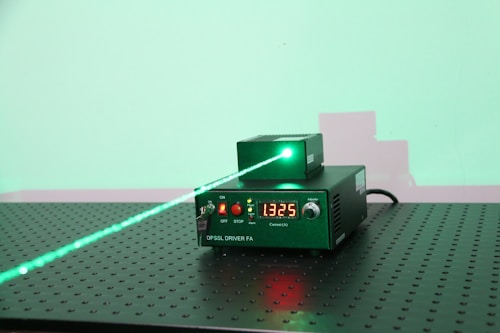 A green laser system placed on a perforated metallic surface. The system consists of a main driver unit labeled 'DPSSL DRIVER FA' and an emitting unit. The front panel of the driver unit has a digital readout displaying the value '1325', buttons, and indicator lights. A visible green laser beam is emitted from the top unit, projecting onto a wall in the background, creating a green spot and a faint shadow of the apparatus.