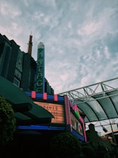 A vintage theater with a neon marquee displaying showtimes under a vast, cloudy sky. The building has art deco architecture and features bright, colorful lights against a darker facade.