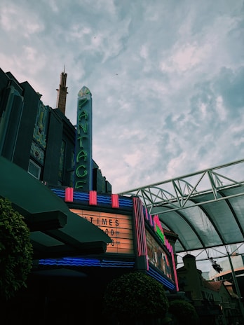 A vintage theater with a neon marquee displaying showtimes under a vast, cloudy sky. The building has art deco architecture and features bright, colorful lights against a darker facade.