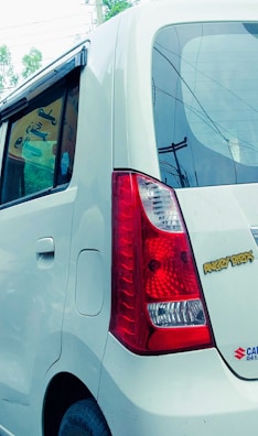 The rear side of a white car with a distinctive red tail light and an 'Angry Birds' sticker. The car window is partially open, revealing some signage inside. Electric poles and wires are visible in the background.