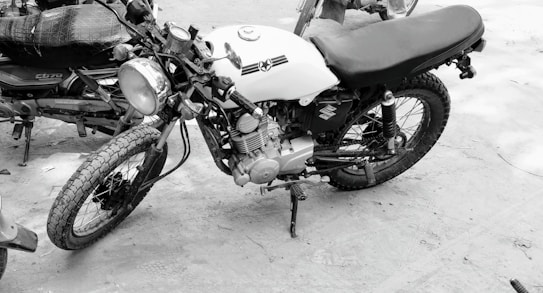 A vintage motorcycle with a sleek black seat and a white fuel tank featuring an emblem. It has a classic round headlight and rugged tires, parked on a sandy surface next to another bike.