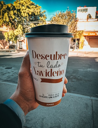A hand is holding a large takeaway coffee cup with festive text written in Spanish. The background shows a street with trees and buildings under a bright blue sky.