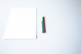 A blank white sheet of paper lies on a smooth, light-colored surface. To the right of the paper, there are four colored pencils aligned together. The pencils are in red, blue, green, and gold colors, and they are neatly positioned.