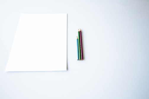 A blank white sheet of paper lies on a smooth, light-colored surface. To the right of the paper, there are four colored pencils aligned together. The pencils are in red, blue, green, and gold colors, and they are neatly positioned.