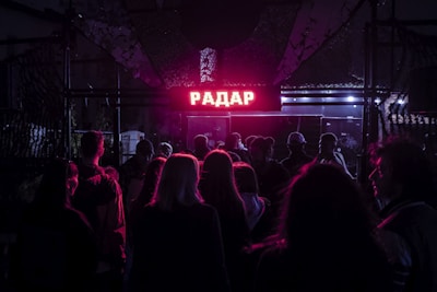 A crowd of people is gathered in front of an entrance marked by a glowing red sign with Cyrillic lettering, suggesting a lively nightlife scene. The area is dimly lit, with a mix of purple and blue ambient lights casting shadows and creating a moody atmosphere.