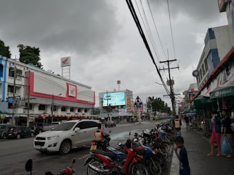 A bustling street scene with several parked motorcycles on one side and various vehicles, including a white SUV, on the road. The sky is overcast with dark clouds, indicating imminent rain. A variety of commercial buildings with colorful signs and advertisements line the street, including a prominent structure with a pink and white facade bearing the Honda logo. People are visible, including a child in the foreground and a person riding a red scooter wearing a safety vest.