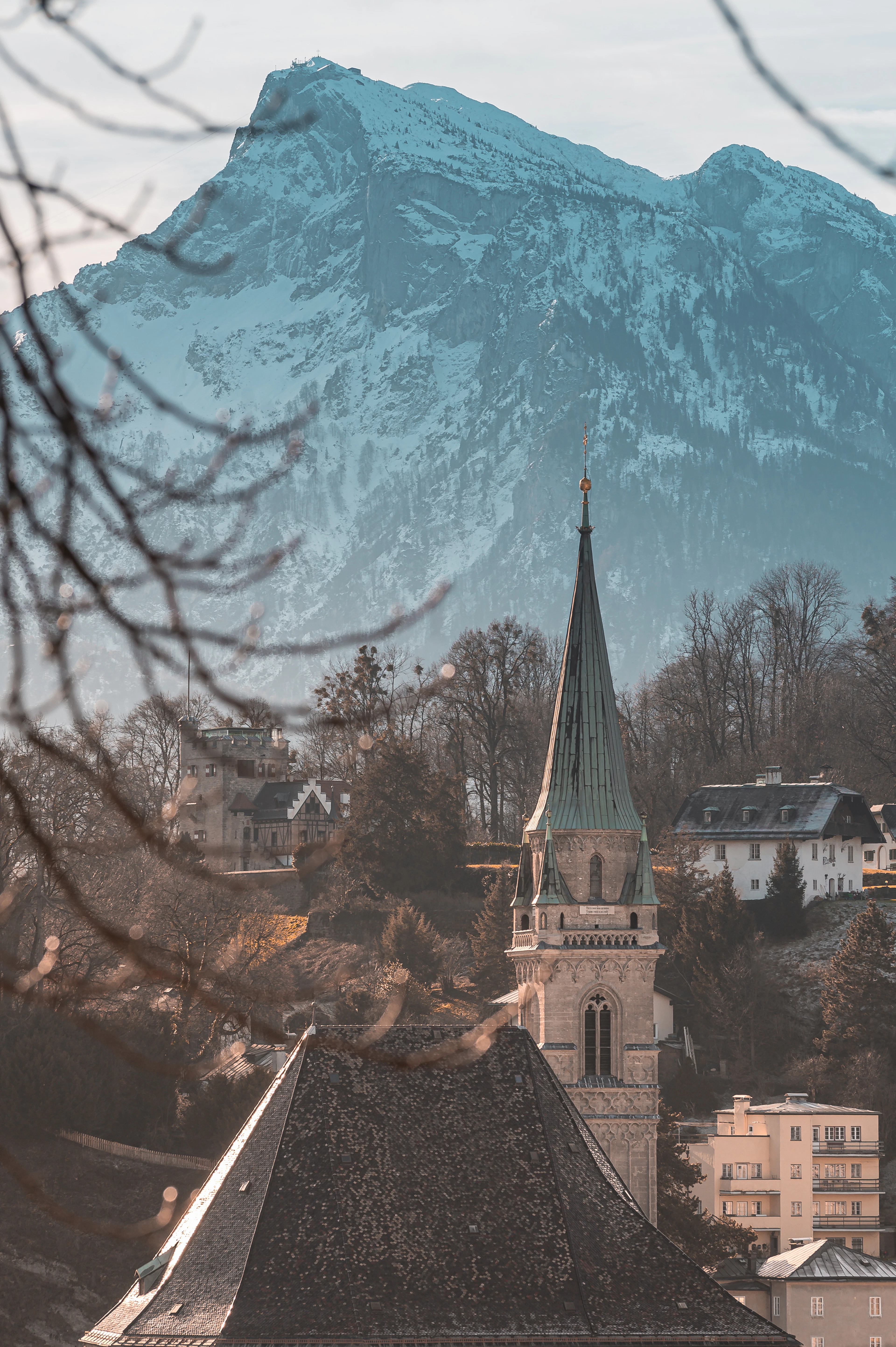 A church spire in Salzburg silhouetted against a snow-capped Alpine peak, framed by bare branches.
