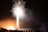 A vibrant fireworks display lights up the night sky with bright white and pink bursts. The silhouettes of people are visible in the foreground, watching the spectacle. The glow from the fireworks illuminates the area with warm light, creating a dynamic and lively scene.