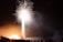 A vibrant fireworks display lights up the night sky with bright white and pink bursts. The silhouettes of people are visible in the foreground, watching the spectacle. The glow from the fireworks illuminates the area with warm light, creating a dynamic and lively scene.