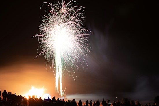 A vibrant fireworks display lights up the night sky with bright white and pink bursts. The silhouettes of people are visible in the foreground, watching the spectacle. The glow from the fireworks illuminates the area with warm light, creating a dynamic and lively scene.