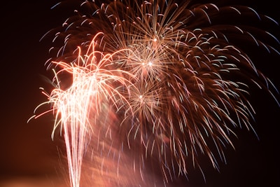 Bright fireworks light up the night sky with bursts of colorful sparks and trails of light. The vibrant display includes a variety of reds, oranges, and whites, creating an explosive and celebratory scene.