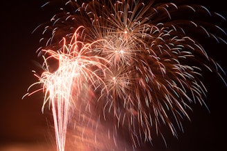 Bright fireworks light up the night sky with bursts of colorful sparks and trails of light. The vibrant display includes a variety of reds, oranges, and whites, creating an explosive and celebratory scene.