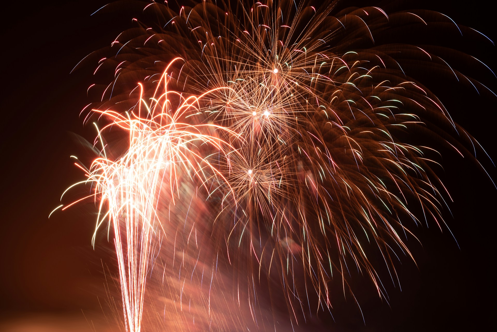 Bright fireworks light up the night sky with bursts of colorful sparks and trails of light. The vibrant display includes a variety of reds, oranges, and whites, creating an explosive and celebratory scene.