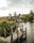 A rustic cottage with a thatched roof sits by a tranquil pond, surrounded by lush greenery and a wooden dock extending into the water. A stone bridge is visible in the distance, along with rolling hills under an overcast sky.