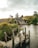 A rustic cottage with a thatched roof sits by a tranquil pond, surrounded by lush greenery and a wooden dock extending into the water. A stone bridge is visible in the distance, along with rolling hills under an overcast sky.