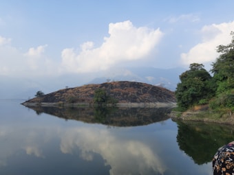 A serene landscape with a calm body of water reflecting a small, shrub-covered hill and a partly cloudy sky. Lush greenery borders the water, creating a peaceful and natural setting.