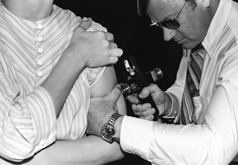 A person in a striped shirt is receiving an injection or vaccination in the arm from another individual wearing glasses and a patterned tie. The second person is focused on the task, holding an injection device close to the other person's arm.