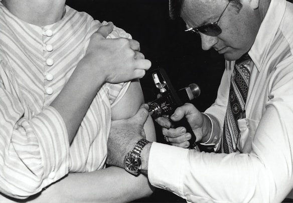 A person in a striped shirt is receiving an injection or vaccination in the arm from another individual wearing glasses and a patterned tie. The second person is focused on the task, holding an injection device close to the other person's arm.