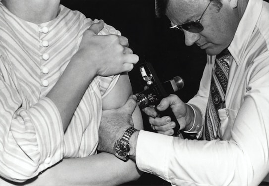 A person in a striped shirt is receiving an injection or vaccination in the arm from another individual wearing glasses and a patterned tie. The second person is focused on the task, holding an injection device close to the other person's arm.