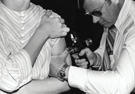 A person in a striped shirt is receiving an injection or vaccination in the arm from another individual wearing glasses and a patterned tie. The second person is focused on the task, holding an injection device close to the other person's arm.