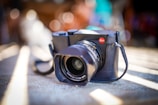 A professional camera with a black body and lens, featuring a prominent red logo on the corner. It is placed on a surface with softly blurred background bokeh effects, highlighting the shiny glass elements of the lens.