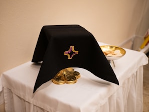 A black cloth with an embroidered cross design covers an ornate gold object, possibly a chalice, on a white tablecloth. To the right, a gold dish holds religious wafers. The setting suggests a religious or ceremonial context.