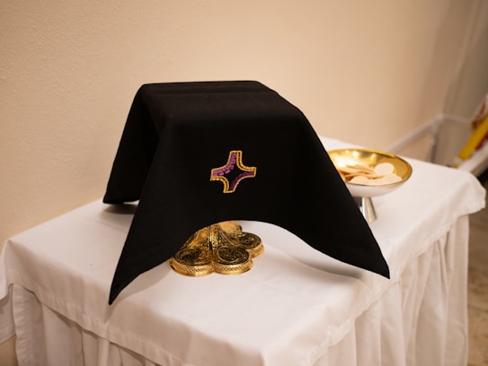 A black cloth with an embroidered cross design covers an ornate gold object, possibly a chalice, on a white tablecloth. To the right, a gold dish holds religious wafers. The setting suggests a religious or ceremonial context.