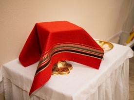 A red cloth with stripes of green and gold covers an ornate gold object on a white draped table. A gold plate with white wafers sits nearby.