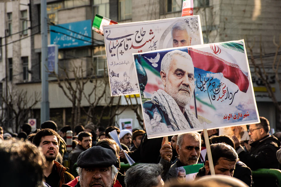 The image depicts a large crowd of people gathered on a street, holding up signs and banners with various messages written in different languages. The most prominent sign is a white banner that reads "مرحبا با ها ألا إنفاء", which translates to "We are united". Other signs include one that says "مرحبا با ها ألا إنفاء" and another with a picture of a man's face. The crowd is diverse, with people standing close together in the foreground and further back in the background.