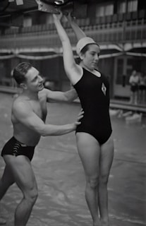 A man and a woman are involved in a gymnastics exercise. The woman is wearing a swimsuit cap and a one-piece swimsuit, hanging onto a bar above her head. The man is supporting her by the waist, ensuring her form and balance. They are in an indoor setting that resembles a swimming pool area.
