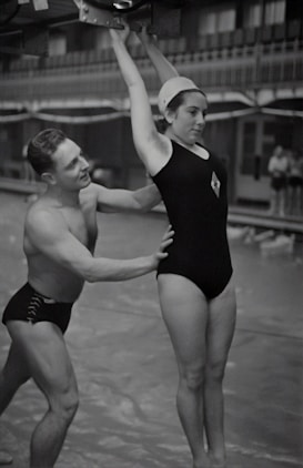 A man and a woman are involved in a gymnastics exercise. The woman is wearing a swimsuit cap and a one-piece swimsuit, hanging onto a bar above her head. The man is supporting her by the waist, ensuring her form and balance. They are in an indoor setting that resembles a swimming pool area.